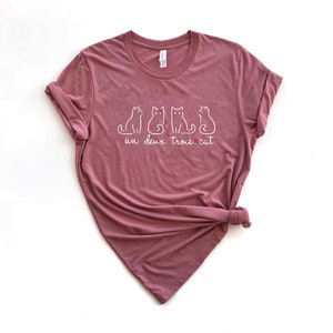 May include: A mauve t-shirt with a white graphic of three cats and the text "un deux trois cat".