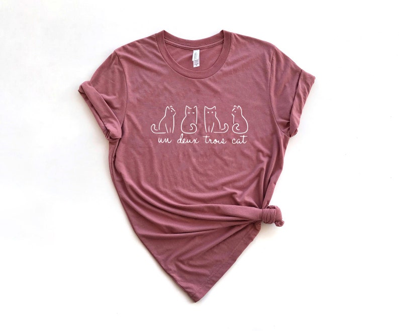 May include: A mauve t-shirt with a white graphic of three cats and the text "un deux trois cat".