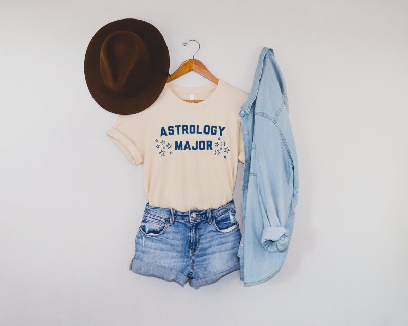 K&ouml;nnte beinhalten: Ein hellbeiges T-Shirt mit dem Text "Astrology Major" in blauer Schrift und kleinen blauen Sternen. Das Shirt h&auml;ngt an einem Holzb&uuml;gel mit einem braunen Hut und einer hellblauen Jeansjacke.