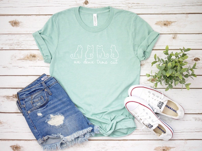 May include: A light green t-shirt with a white graphic of three cats and the text "un deux trois cat". The shirt is paired with denim shorts and white sneakers.