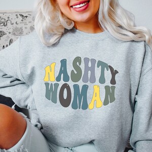 Könnte beinhalten: Ein graues Sweatshirt mit dem Text "Nasty Woman" in einer Retro-Schriftart in verschiedenen Farben. Die Buchstaben sind in Gelb, Grün und Blau umrandet.
