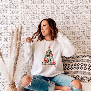 May include: White sweatshirt featuring a Christmas tree design made of dogs, with the text "MERRY WOOFMAS" in red and green. The sweatshirt is worn with ripped blue jeans.