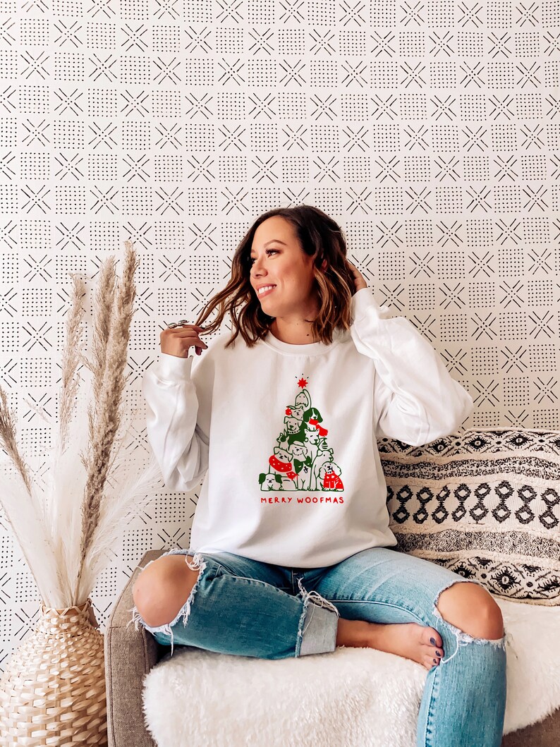 May include: White sweatshirt featuring a Christmas tree design made of dogs, with the text "MERRY WOOFMAS" in red and green. The sweatshirt is worn with ripped blue jeans.