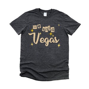 May include: A dark heather grey t-shirt with the text "We said Vegas" in a tan color with gold stars.