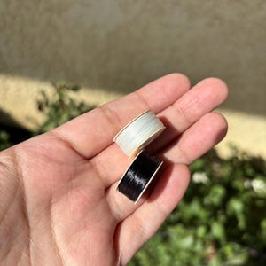 May include: Two spools of thread, one white and one black, are held in a hand. The spools are small, with the thread neatly wound around cardboard cores. The background is blurred, with hints of green foliage and a neutral wall.