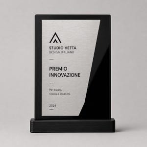 Può includere: Una targa premio rettangolare con cornice e base nere, dotata di un pannello centrale in metallo argento spazzolato. Il testo riporta STUDIO VETTA DESIGN ITALIANO, PREMIO INNOVAZIONE, Per visione, ricerca e creatività e l'anno 2024 in nero.