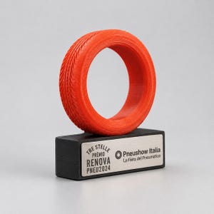 Könnte beinhalten: Eine orangefarbene, reifenförmige Auszeichnung steht auf einem schwarzen, rechteckigen Sockel mit einem silbernen Namensschild. Das Namensschild trägt italienischen Text, darunter "TRE STELLE PREMIO RENOVA PNEU2024" und "Pneushow Italia La Fiera del Pneumatico."