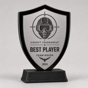 Può includere: Un trofeo a forma di scudo con bordo nero e piastra centrale in argento spazzolato. Presenta un grafico di un casco tattico all'interno di un mirino, con il testo AIRSOFT TOURNAMENT, BEST PLAYER, TEAM RAVEN e 2024 inciso sulla parte anteriore.