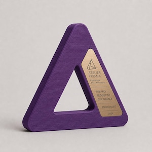 Könnte beinhalten: Eine dreieckige violette Auszeichnung mit strukturierter Oberfläche und einer goldfarbenen Metalleinlage. Die Einlage zeigt ein geometrisches Logo und den Text: ATELIER PRISMA, Associazione Arte e Architettura, PREMIO PROGETTO CULTURALE, STUDIO LUCE, 2024.