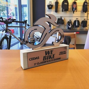 Può includere: Un trofeo a forma di bicicletta color argento con le parole "WE BIKE" e "3° CLASSIFICATO DONNE" su una base di legno. Il trofeo è su un tavolo di legno, con biciclette e attrezzatura da ciclismo visibili sullo sfondo.