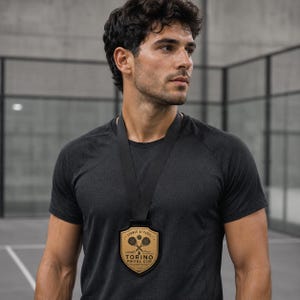 Può includere: Una medaglia nera con un inserto centrale color oro a forma di scudo appesa a un nastro nero. La medaglia riporta la scritta TORINO PADEL CUP, un'illustrazione di due racchette da padel incrociate e una pallina, con l'anno 2020 stampato sotto.