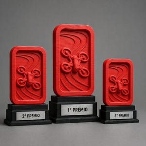 Peut inclure: Trois trophées rectangulaires rouges avec un relief de drone et une texture de fond ondulée. Chaque trophée repose sur une base noire avec une plaque métallique argentée. Les plaques portent les inscriptions 1° PREMIO, 2° PREMIO et 3° PREMIO.