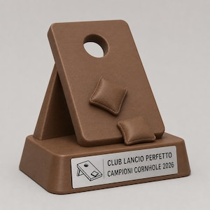 May include: A brown cornhole trophy featuring a miniature board with a circular hole and two small bean bags attached to the surface. The base includes a silver plaque with a cornhole board icon and the text CLUB LANCIO PERFETTO CAMPIONI CORNHOLE 2026.