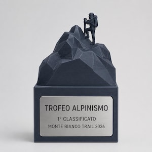Może przedstawiać: Ciemnoniebieskie trofeum przedstawiające geometryczny szczyt górski z małą figurką wspinacza na górze. Prostokątna podstawa posiada srebrną metalową tabliczkę z czarnym napisem: TROFEO ALPINISMO, 1° CLASSIFICATO, MONTE BIANCO TRAIL 2026.