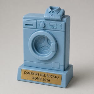 Puede incluir: Un trofeo azul claro con forma de lavadora y una camisa doblada encima. La base presenta una placa dorada con el texto negro "CAMPIONE DEL BUCATO NOME 2026" grabado en la parte frontal, destacando la puerta circular del electrodoméstico.