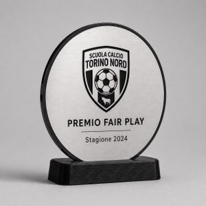 Puede incluir: Una placa de premio circular con acabado en plata cepillada y borde negro, montada sobre una base rectangular negra. El centro presenta un logotipo de escudo con un icono de balón de fútbol y un toro, con el texto SCUOLA CALCIO TORINO NORD, PREMIO FAIR PLAY, Stagione 2024.