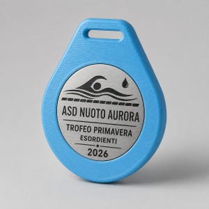 Könnte beinhalten: Eine hellblaue, tropfenförmige Kunststoffmedaille mit einem kreisförmigen, silberfarbenen Metalleinsatz. Der Einsatz zeigt eine schwarze Grafik eines Schwimmers, einen Wassertropfen sowie den Text ASD NUOTO AURORA, TROFEO PRIMAVERA ESORDIENTI, 2026.