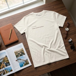 May include: Off-white t-shirt with the text "Yo lo buscaba... y Él me encontró primero" in dark brown. The shirt is on a wooden surface with a planner, pen, magazine, watch, and sunglasses.