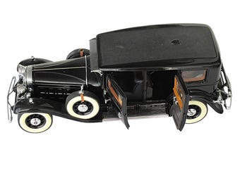 Op de afbeelding: Een zwart vintage automodel met open deuren, witwandbanden en een zwart dak. De auto heeft chromen accenten en een reservewiel aan de achterkant. Het model staat tegen een witte achtergrond.