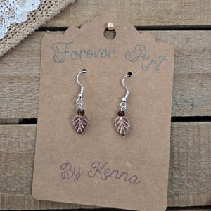 May include: A pair of leaf-shaped earrings with silver hooks and small brown beads. The earrings are displayed on a brown card with the text "Forever Art By Kenna". The background is a wooden surface.