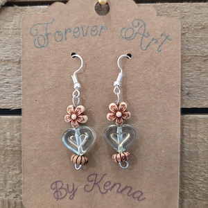 May include: A pair of dangle earrings with silver hooks. Each earring features a clear heart-shaped bead, a small flower bead, and a decorative bead. The earrings are displayed on a brown card with the text "Forever Art By Kenna."