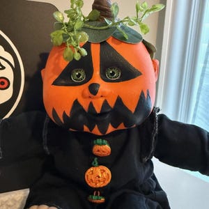 Puede incluir: Una muñeca con cabeza de calabaza naranja redonda, ojos triangulares pintados de negro y boca dentada. La muñeca viste un conjunto negro de manga larga adornado con tres pequeños botones en forma de calabaza y hojas de plástico verdes.