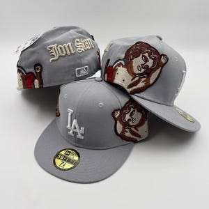 May include: Three gray baseball caps with embroidered designs. One cap features "Jon Stan" in white script, another displays the LA logo, and all have a cherub patch. The caps are 59FIFTY fitted hats.