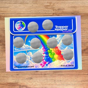 May include: A blue Trapper Keeper notebook featuring a rainbow and heart graphic design. The cover includes ten circular silver scratch-off areas, the text Trapper Keeper NOTEBOOK, DreamItSaveIt, and Save $45 printed in white and black lettering.