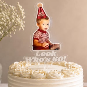 May include: A round white frosted cake with gold sprinkles at the base, topped with a clear acrylic cake topper featuring a photograph of a child wearing a red party hat and the white text Look Who's 60! printed on the base of the topper.