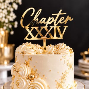Könnte beinhalten: Ein goldfarbener Acryl-Tortenstecker mit dem Wort Chapter in Schreibschrift über der römischen Ziffer XXXIV in fetter Blockschrift, montiert auf einem dünnen Stiel, platziert auf einer weiß glasierten Torte mit Blattgold und Perlen verziert.