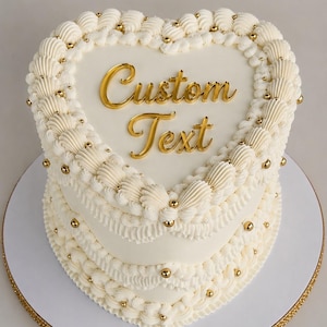 May include: A heart-shaped cake with smooth white frosting, decorated with intricate white piped borders and small gold spherical sprinkles. The top of the cake features the gold-colored text "Custom Text" in a cursive script font.