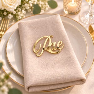 May include: A gold-colored acrylic name tag featuring the name Rae in a cursive script font, placed on a folded beige linen napkin resting on a white ceramic plate with a gold rim, creating a decorative table setting for a formal event.