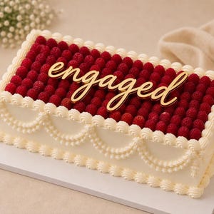 May include: A rectangular cake covered in fresh red raspberries with white buttercream frosting piped in decorative swags and borders. A gold-coloured acrylic cake topper in cursive script reads the word engaged, centred on top of the fruit layer.