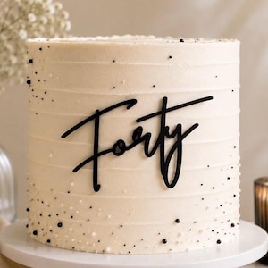 May include: A cylindrical cake with smooth off-white frosting featuring horizontal ridges. The cake is decorated with small black and white sprinkles scattered across the surface and a black script cake topper that reads the word Forty.