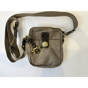 Peut inclure: Sac bandoulière taupe avec une bandoulière réglable. Le sac est doté d'une poche avant, d'un détail en cuir noir et d'un emblème circulaire doré. Un charm en forme de singe est suspendu à la fermeture éclair.
