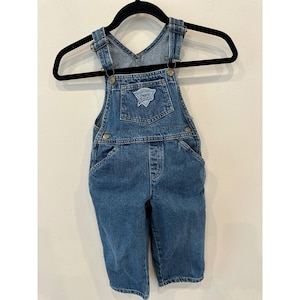 May include: Blue denim overalls with a Guess logo patch on the chest. The overalls have adjustable straps, pockets, and metal hardware. The garment is hanging on a black hanger.