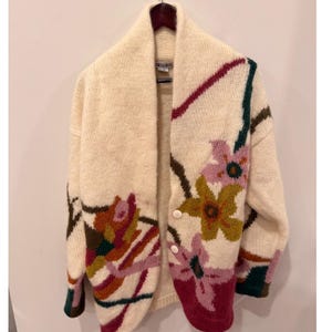 May include: A cream-colored knit cardigan featuring a shawl collar and two white buttons. The garment is decorated with a large-scale floral pattern in shades of magenta, mustard yellow, dark green, and brown, extending across the front and sleeves.