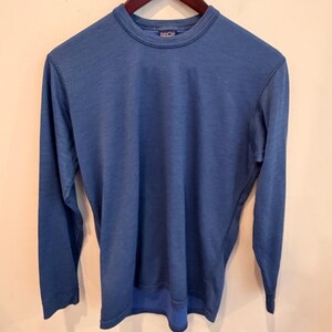 Puede incluir: Una camiseta de manga larga con cuello redondo en un color azul medio sólido. La prenda presenta un cuello acanalado, un dobladillo recto y una etiqueta de marca visible en el interior del cuello. La tela tiene una textura fina y sutil.