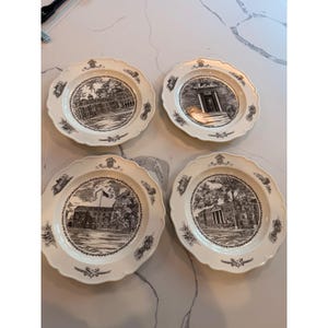 May include: Four white decorative plates with scalloped edges. Each plate features a black and white illustration of a building or architectural scene. The plates have a decorative border with floral and scroll elements.