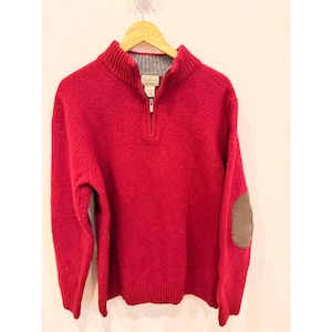 Może przedstawiać: Czerwony wełniany sweter z zapięciem na ćwierć zamka i szarym wewnętrznym kołnierzem. Sweter ma brązową naszywkę na łokciu na prawym rękawie. Sweter wisi na drewnianym wieszaku.