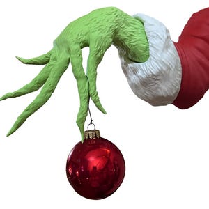 Puede incluir: Una mano verde texturizada sosteniendo una brillante bola de Navidad roja. La mano está unida a una manga roja con ribete de piel sintética blanca, lo que sugiere un tema festivo. La bola tiene una tapa dorada.