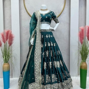 May include: A teal-colored, embroidered lehenga choli set displayed on a mannequin. The set includes a fitted top, a flowing skirt, and a sheer dupatta. The outfit features intricate gold embroidery and a matching gold necklace.