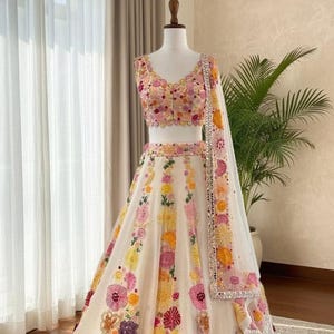 Puede incluir: Conjunto lehenga choli color crema con bordados florales. El conjunto presenta un top corto y una falda larga, ambos adornados con patrones florales coloridos. Una dupatta a juego con un borde decorativo completa el look. El atuendo se exhibe en un maniquí.