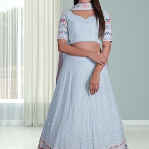 May include: A pale blue lehenga choli ensemble featuring intricate embroidery. The outfit comprises a short-sleeved top, a flowing skirt, and a coordinating scarf. Floral embroidery adorns the sleeves and the skirt's hem.