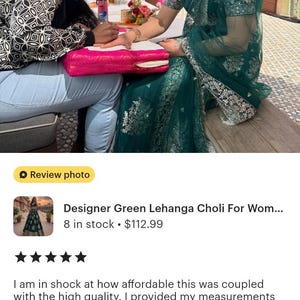 Może przedstawiać: Kobieta w zielonym Lehenga Choli z misternym haftem, podczas gdy na jej dłoń nakładana jest henna. Scena rozgrywa się na zewnątrz, udekorowana r&oacute;żem i pomarańczem, prawdopodobnie z okazji uroczystości.
