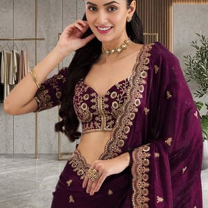 Peut inclure: Un sari prune avec broderie dor&eacute;e. Le sari pr&eacute;sente un chemisier assorti avec des d&eacute;tails dor&eacute;s complexes et un d&eacute;collet&eacute; plongeant. L'ensemble comprend un ras du cou, des boucles d'oreilles et une grosse bague dor&eacute;s. L'arri&egrave;re-plan montre un portant et une plante.