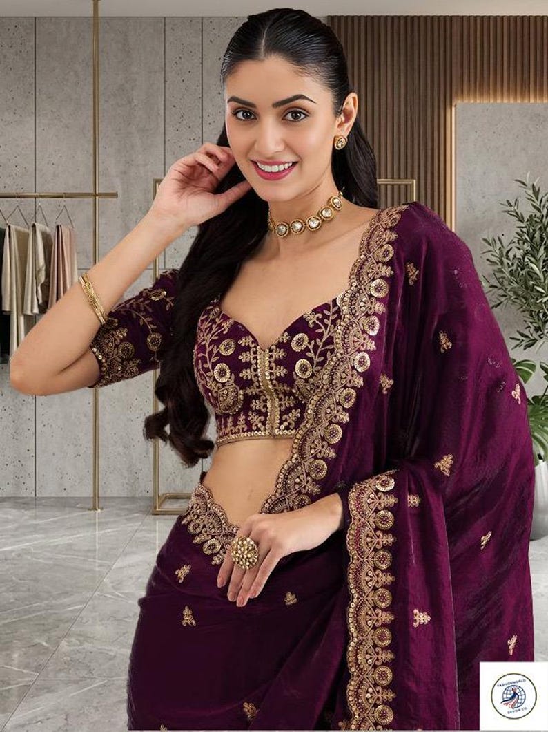Peut inclure: Un sari prune avec broderie dor&eacute;e. Le sari pr&eacute;sente un chemisier assorti avec des d&eacute;tails dor&eacute;s complexes et un d&eacute;collet&eacute; plongeant. L'ensemble comprend un ras du cou, des boucles d'oreilles et une grosse bague dor&eacute;s. L'arri&egrave;re-plan montre un portant et une plante.