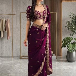 Peut inclure: Une femme porte un sari violet fonc&eacute; avec des broderies dor&eacute;es. Le sari comprend un chemisier assorti avec des manches courtes et des d&eacute;tails dor&eacute;s complexes. Le tissu semble &ecirc;tre du velours, et le sari est drap&eacute; avec &eacute;l&eacute;gance. Le style g&eacute;n&eacute;ral est traditionnel et formel.