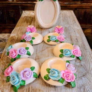May include: A collection of decorative candle holders and a picture frame. The candle holders are beige with colorful rose accents in pink, blue, and purple. The picture frame is oval-shaped and adorned with similar floral details.