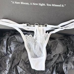 May include: White lingerie set with a delicate lace design. The thong features a decorative silver chain and ring detail. The set is displayed in a black box with the text "A New Bloom, A New Sight. You Missed It."